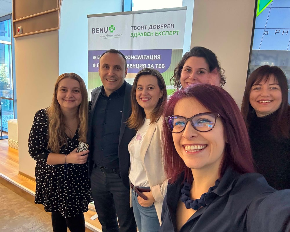 Група усмихнати хора позират за селфи пред банер на BENU с надпис „Твоят доверен здравен експерт“.
